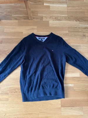 Mörkblå stickad tröja Tommy Hilfiger - Snygg mörkblå stickad tröja från Tommy Hilfiger med V-ringning och diskret logga på bröstet. Tröjan har långa ärmar och ribbade muddar vid ärmslut och nederkant. Perfekt till jeans eller chinos för en clean look.
