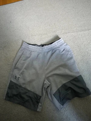 Grå sportshorts från Under Armour - Snygga grå sportshorts från Under Armour med mörkare grå paneler längst ner på benen. Shortsen har elastisk midja och två sidofickor. Perfekta för träning eller chill. Materialet känns mjukt och stretchigt.