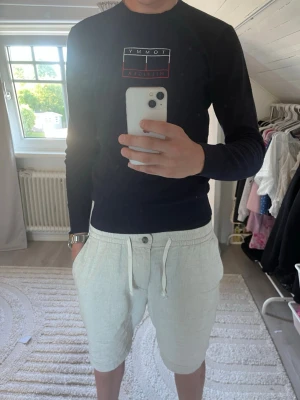 Blå tröja av Tommy Hilfiger - Ljusbeige shorts med normal passform och snörning i midjan. Shortsens material ser ut att vara linne eller bomull, vilket gör dem luftiga och bekväma. De har knappar framtill och två sidofickor för en avslappnad stil.