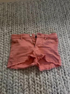 Korallrosa jeansshorts från Gant - Snygga korallrosa jeansshorts från Gant med fransig kant och klassisk femficksdesign. Shortsen har knapp och dragkedja framtill samt bälteshällor. Perfekta för varma dagar och ger en avslappnad vibe. Små i storleken