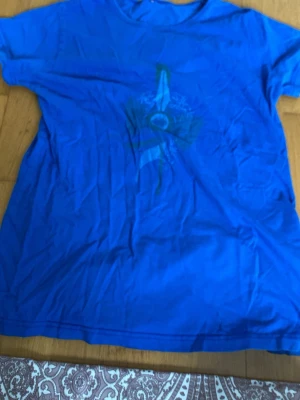 Blå t-shirt med tryck - En blå t-shirt med ett abstrakt tryck i grönt och vitt på bröstet. Klassisk rund halsringning och korta ärmar. Perfekt för en avslappnad stil och enkel att matcha med jeans eller shorts.