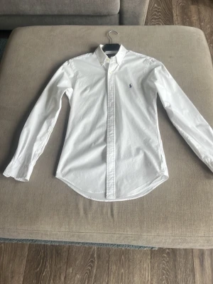 Vit slim fit skjorta Polo Ralph Lauren - Klassisk vit skjorta från Polo Ralph Lauren i slim fit-modell. Skjortan har lång ärm, knappar framtill och den ikoniska broderade loggan på bröstet. Tillverkad i bomull och har en tidlös, clean look som passar till allt.