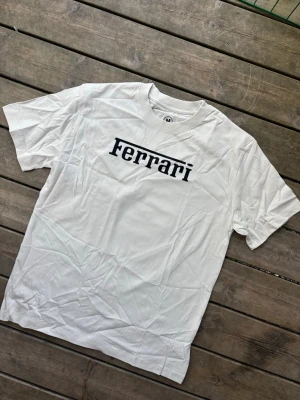 Vit Ferrari t-shirt med tryck Charles Leclerc  - Snygg vit t-shirt från Ferrari med svart logga på bröstet och ett stort färgglatt motiv av en Formel 1-förare på ryggen. Klassisk rund hals och korta ärmar. Perfekt för dig som gillar motorsport och vill sticka ut med en unik design.