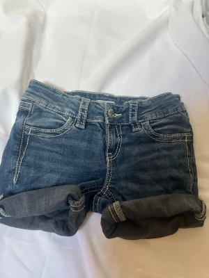 Blå jeansshorts med rå kant - Säljer ett par klassiska blå jeansshorts med rå kant och kontrastsömmar. Shortsen har fem fickor, bälteshällor och kan bäras både upprullade och nedvikta för olika stil. Perfekta till sommaren och lätta att matcha med allt. (Kommer från Gina) storlek:146