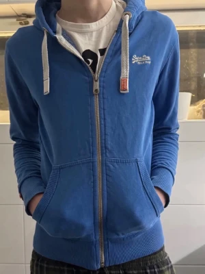 Blå zip-hoodie från Superdry - Snygg blå hoodie från Superdry med dragkedja, vita snören i huvan och två stora fickor framtill. Märkeslogga broderad på bröstet och detaljer i vitt och orange. Perfekt för en avslappnad och cool stil.