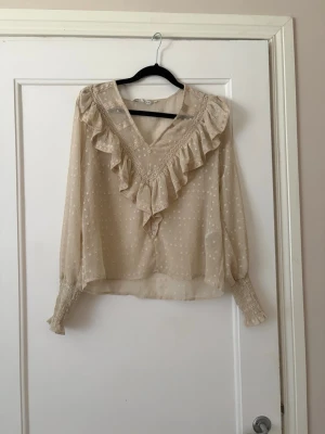 Beige blus med volang och prickar - Söt beige blus från only i storlek S🤍Blusen har transparent material och små utåtstående prickar. Blusen har en djup V-ringning med bred volangdetalj framtill och långa ärmar med smock vid handlederna. Perfekt för en trendig och romantisk look. Endast använd max 5ggr🤍