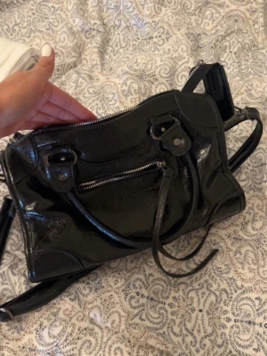 Svart lackad handväska med dragkedja - Snygg svart handväska i glansigt lackmaterial med både handtag och axelrem. Väskan har en framficka med dragkedja och dekorativa remmar. Perfekt för dig som gillar en edgy och trendig look.