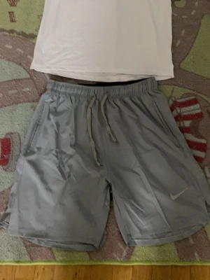 Grå Nike shorts med snörning - Snygga grå shorts från Nike med elastisk midja och snörning för skön passform. Shortsen har sidofickor och är perfekta för träning eller chill. Klassisk Nike-logga på benet och tillverkade i lätt syntetmaterial.
