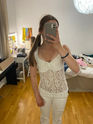 Vit spetslinne med knyt framtill - Snyggt vitt spetslinne med v-ringning och knyt framtill. Linnet är ärmlöst och har ett genombrutet blommigt spetsmönster som ger en romantisk vibe. Perfekt att styla med jeans eller kjol för en trendig look.