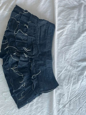 Mörkblå volangkjol från Bershka - Kort volangkjol från Bershka i mörkblå denim med råa kanter och dekorativa trådar. Kjolen har dragkedja i sidan och en tight passform i midjan. Perfekt för dig som gillar en edgy och trendig stil.