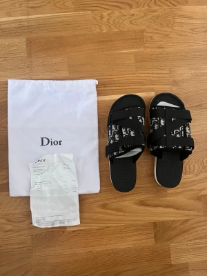 Svarta sandaler med logga - Säljer ett par svarta sandaler från med vita detaljer och tydlig -logga på remmarna. Sandalerna har två breda justerbara remmar i textil och en vit sula med märkning undertill. Perfekta för en trendig och avslappnad stil.