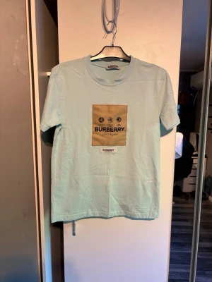 Ljusblå Burberry t-shirt med tryck - Snygg ljusblå t-shirt från Burberry med klassiskt tryck på bröstet i beige och svart. T-shirten har rund halsringning och korta ärmar. Perfekt för dig som gillar stilrena märkesplagg med en modern touch.
