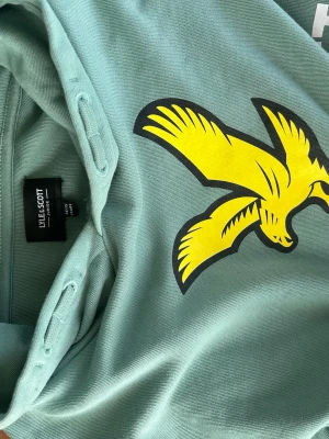 Grön hoodie från Lyle & Scott - Säljer en snygg hoodie från Lyle & Scott i en stilren grön färg med klassisk gul logga framtill. Bekväm passform och mjukt material som passar perfekt till vardags. Nästan aldrig använd och hyfsat ny.  ✅ Bra skick ✅ Stor framficka ✅ Klassisk och tidlös design ✅ Perfekt för både vår, sommar och höstkvällar
