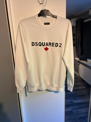 Vit Dsquared2 sweatshirt - Vit sweatshirt från Dsquared2 med svart logotyp och rött lönnlöv broderat på bröstet. Tröjan har rund hals, långa ärmar och ribbade muddar. Tillverkad i mjukt material som ger en clean och modern look.