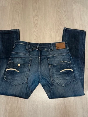 G-Star Raw mörkblå jeans - Säljer ett par mörkblå jeans från G-Star Raw med coola detaljer på bakfickorna och läderpatch vid linningen. Jeansen har raka ben och slitningar som ger en snygg, använd look. Materialet är klassisk denim och passformen är relaxed.