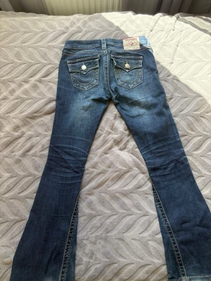 True Religion Jeans Joey ”low rise flare” - Säljer ett par blå bootcut jeans från True Religion med snygga kontrastsömmar och dekorativa fickor bak med vita knappar. DEFEKTER PÅ BILD 4,5,6 OCH DÄRAV PRISET!! Defekterna går säkert att fixa genom att sy några stygn och defekterna syns inte när man har på sig jeansen❤️❤️Jeansen har en klassisk femficksmodell och är tillverkade i stretchig denim för skön passform. Pris går att diskutera!💞