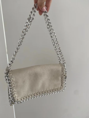 Beige axelremsväska med kedja - Stilren beige handväska i mockaimitation med silverfärgade kedjedetaljer runt kanterna och som axelrem. Väskan har en rektangulär form och coola sömmar längs kanten som ger en trendig look. Perfekt accessoar för att lyfta din outfit.