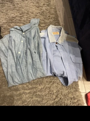 Två blå skjortor, casual stil - Säljer två blå skjortor i avslappnad stil. Ena skjortan är ljusblå med vita knappar och liten broderad detalj på fickan. Den andra är ljusblå med vit krage och fickor framtill, från Samsøe Samsøe. Perfekta för en chill look.