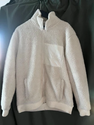 Vit teddyjacka Peak Performance - Mysig vit teddyjacka från Peak Performance med hög krage, hel dragkedja och en bröstficka med dragkedja. Jackan har två sidofickor och ribbade muddar vid ärmslut och nederkant. Perfekt för kalla dagar och riktigt skön att bära.