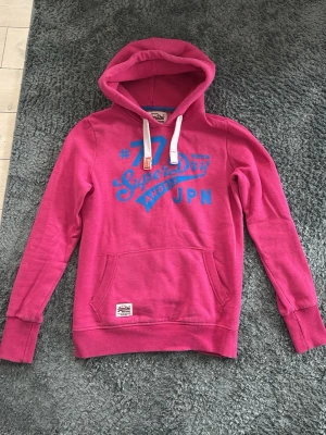 Rosa/röd Superdry hoodie med tryck - Snygg rosa hoodie från Superdry med blått tryck fram och stort nummer 77 på ryggen. Klassisk känguruficka, vita dragsnören i huvan och ribbade muddar. Perfekt för dig som gillar sportig streetstyle och vill sticka ut lite extra. Rätt liten i storlek så hade sagt S.