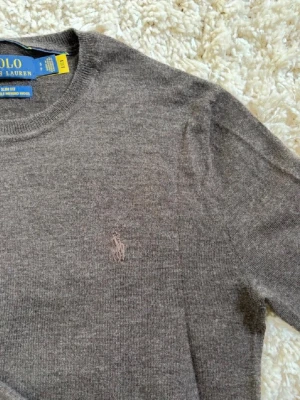 Brun stickad tröja från Polo Ralph Lauren - Klassisk brun stickad tröja från Polo Ralph Lauren i mjuk merinoull. Tröjan har rund halsringning, långa ärmar och diskret broderad logga på bröstet. Perfekt till både jeans och chinos för en stilren look.