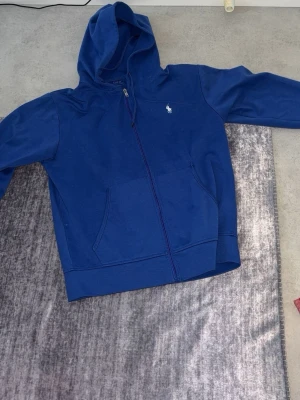 Blå hoodie från Polo Ralph Lauren - Blå hoodie med dragkedja från Polo Ralph Lauren. Klassisk design med vit broderad logga på bröstet, kängurufickor och huva. Tillverkad i mjuk bomullsblandning som är skön att ha på sig. Perfekt för en avslappnad och stilren look.