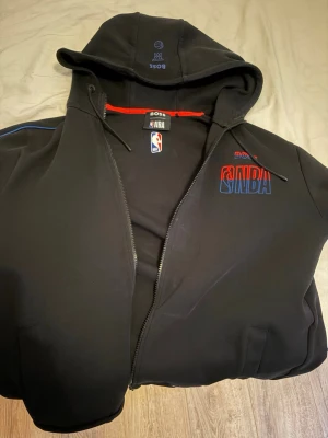 Svart NBA hoodie från BOSS - Cool svart hoodie från BOSS x NBA med dragkedja och huva. NBA-logga och BOSS-tryck på bröstet, samt snygga detaljer i rött och blått. Perfekt för dig som gillar basket och streetwear. Materialet känns mjukt och bekvämt, med snören i huvan. Har gått ner i vikt så passar ej längre