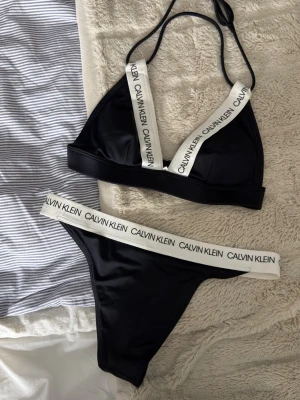 Svart bikini från Calvin Klein - Snygg svart bikini från Calvin Klein i storlek xs med vita resårband där loggan syns tydligt. Överdelen har trekantskupor och smala axelband som knyts i nacken, samt spänne i ryggen. Underdelen har en klassisk passform med bred resår i midjan. Klassiker till sommaren. Säljer då den hängt i garderoben och inte används💞(inga defekter)