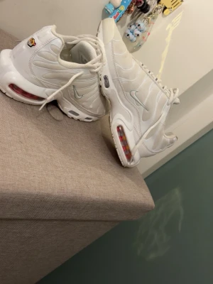 Nike Air Max Plus vita sneakers - Nike Air Max Plus sneakers i helvitt med ikoniska vågiga detaljer och synlig Air-enhet i sulan. Skorna har mesh- och syntetmaterial, vita skosnören och små röda detaljer i sulan. Perfekta för dig som gillar sportig och clean stil.
