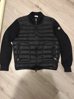 Svart pufferjacka från Moncler - Snygg svart pufferjacka från Moncler med quiltad front och släta ärmar. Jackan har två dragkedjeförsedda fickor och en hög krage. Moncler-logga på vänster ärm. Perfekt för kalla vinterdagar och riktigt trendig look. 