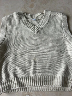 Ljus stickad väst med v-ringning - En stilren ljus stickad väst med v-ringad hals och bred ribbad kant nedtill. Perfekt att slänga över en skjorta eller t-shirt för en clean look. Västen har en enkel och klassisk design utan mönster eller detaljer.