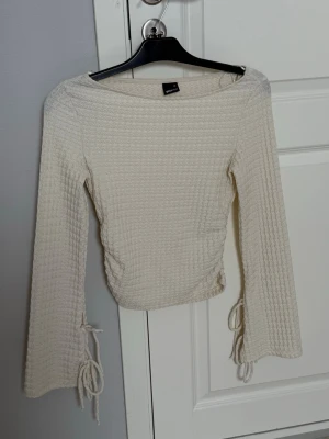 Beige ribbad topp med snörning - Trendig beige ribbad topp från Gina Tricot med långa ärmar och snörningsdetaljer vid ärmsluten. Toppen har en croppad passform och är tillverkad i mjukt material som känns skönt mot huden. Perfekt för att lyfta din outfit.