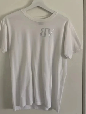 Vit t-shirt från bvaldi med strasstenar - Snygg vit t-shirt från bvaldi med korta ärmar. Framtill finns ett glittrigt VB-tryck i strasstenar och på ryggen står det bvALDI i blå och silverfärgade stenar. Perfekt för dig som gillar stilrena plagg med en twist.