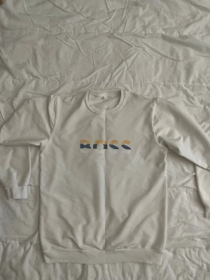 Vit sweatshirt från BOSS - Snygg vit sweatshirt från BOSS med tryckt logga i blått, gult och vitt på bröstet. Tröjan har rund halsringning, långa ärmar och ribbade muddar vid ärmslut och nederkant. Perfekt för en clean och stilren look.