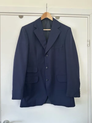 Mörkblå klassisk kavaj från Ströms i storlek S - Stilren mörkblå / navy kavaj / blazer från anrika kostymtillverkaren Ströms Harrington de Luxe i storlek S. Klassisk helfodrad modell  knappar framtill och fickor med lock. Stiliga dekorativa knappar vid ärmsluten. Praktisk sprund baktill för ökad rörlighet. 100% ull, så bra material som andas. Tidlös välkonstruerad klassiker som kommer hålla säsong efter säsong. Nypris 4999:- . Mycket gott skick, som ny!