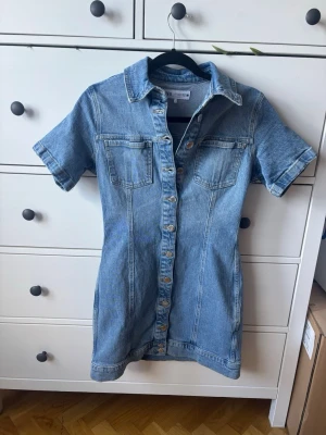 Kort jeansklänning från Zara - Säljer en snygg kort jeansklänning från Zara med knappar framtill och två bröstfickor. Klänningen har korta ärmar, klassisk skjortkrage och är tillverkad i blått jeanstyg. Perfekt för en trendig och avslappnad look.