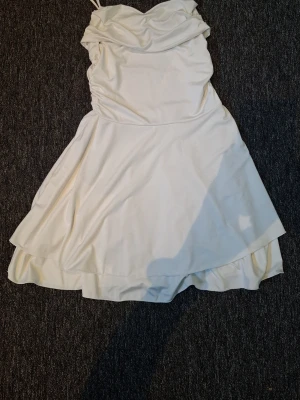 Vit kort klänning med tunna axelband - Säljer en vit kort klänning med tunna axelband och draperad urringning. Klänningen har en lätt utsvängd kjol och sitter figurnära upptill. Perfekt för sommarens alla tillfällen!