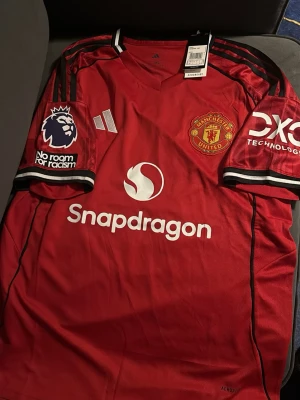 Manchester United fotbollströja Fernandes - Röd Manchester United matchtröja med svarta och vita detaljer. Tröjan har klubbmärke, Premier League-märke och sponsorloggor på bröst och ärmar. På ryggen står det B. Fernandes och nummer 8. Tillverkad i lätt och ventilerande material. Storlek L och har aldrig använts 