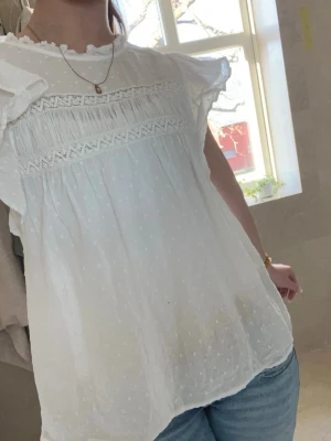Vit blus med volang och spetsdetaljer - Söt vit blus med volangärmar och spetsdetaljer över bröstet. Blusen har diskret prickigt mönster i tyget och en lös, luftig passform. Det finns en liten liten svart prick på den som jag inte riktigt har fått bort än när jag tvättat, den kan gå bort om man andvänder något starkare vilket jag inte testat. Annars så ser man inte den lilla fläcken alls tydligt, man kan se den lite i bilderna om du kollar noga. Säljer för att jag ändrat min stil mycket och jag andvänder inte blusar lika ofta! 💞