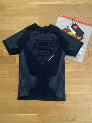 Svart Superman t-shirt från YoungLA - Cool svart sport-t-shirt från YoungLA med stort Superman-tryck på bröstet. Tröjan har korta ärmar och gråmönstrade paneler på sidorna och ärmarna. Perfekt för gymmet eller träning. Materialet är stretchigt och andas bra.