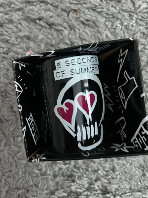 5 Seconds of Summer svart mugg - Cool svart mugg med 5 Seconds of Summer-tryck och en vit dödskalle med hjärtformade ögon i rosa. Muggen levereras i en matchande svart kartong med vita grafiska detaljer. Perfekt för fans av bandet eller som samlarpryl. Helt ny.