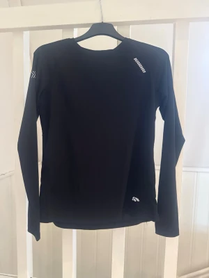 Svart långärmad träningströja - Svart långärmad träningströja från karhu med diskreta vita logotyper på bröstet och ärmen. Tillverkad i ett stretchigt funktionsmaterial som andas, perfekt för löpning eller gym. Enkel och stilren design med rund hals.