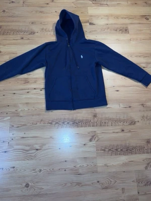 Mörkblå hoodie från Polo Ralph Lauren - Säljer en mörkblå hoodie med dragkedja från Polo Ralph Lauren. Hoodien har huva med snören, två stora fickor framtill och den klassiska vita Polo-loggan på bröstet. Perfekt för en avslappnad och cool look.