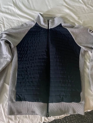 Grå och svart jacka med quiltad front från colmar - Snygg zipjacka från colmar med quiltad svart front och gråa ärmar samt rygg. Hög krage och ribbade muddar. Perfekt för lager-på-lager och chill stil. Märke på vänster ärm för extra detalj.