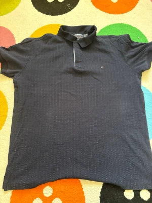 Mörkblå piké från Tommy Hilfiger - Snygg mörkblå pikétröja från Tommy Hilfiger med diskret prickigt mönster och klassisk krage. Tröjan har korta ärmar och liten logga broderad på bröstet. Perfekt till jeans eller shorts för en clean look.