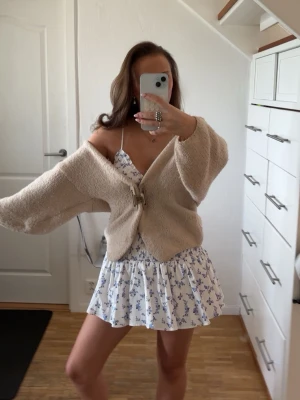 Vit blommig kortklänning med volang - Söt vit kortklänning med blått blommönster och volang nedtill. Klänningen har smala axelband och smockad överdel som ger en bekväm passform. Perfekt för varma dagar och somriga tillfällen.