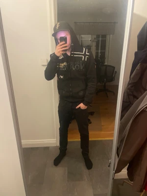 Svart Geographical norway hoodie med tryck - Säljer en svart hoodie från Geographical norway med vita grafiska tryck och detaljer på bröstet och axlarna. Hoodien har dragkedja, fickor framtill och en huva. Perfekt för dig som gillar streetwear och vill ha en snygg och bekväm look.