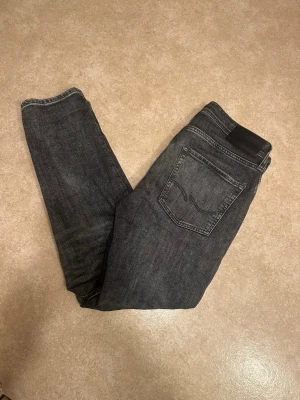 Gråa jeans från Jack & Jones  - Säljer ett par gråa jeans från Jack & Jones 29/30 med slitningar på låret och knät. Jeansen har klassisk femficksmodell, normal midja och är tillverkade i stretchigt denimtyg för extra komfort. Perfekta för dig som gillar en avslappnad men trendig look.