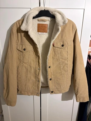 Beige manchesterjacka från Levi's - Snygg beige jeansjacka i manchester från Levi's med mysigt vitt teddyfoder. Klassisk modell med två bröstfickor, knappar framtill och krage. Perfekt för dig som vill ha en cool och tidlös look med extra komfort.