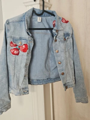 Ljusblå jeansjacka med broderade rosor - Snygg ljusblå jeansjacka från H&M med broderade röda rosor på axeln och bröstfickan. Klassisk modell med knappar framtill och två bröstfickor. Perfekt för dig som vill sticka ut lite extra.